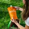 Person holding an orange Sunday “Heat Defense” lawn treatment pouch above an open cardboard box on grass.