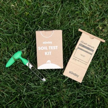 Sunday soil test kit items arranged on grass: shipping box, soil sample bag, and green soil corer.