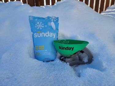 Pet Safe Ice Melt and Sunday Spreader in snow bank alongside yard fence.
