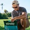 Person kneeling on a sunny lawn opening a green Sunday “Lawn Strong” fertilizer bag.