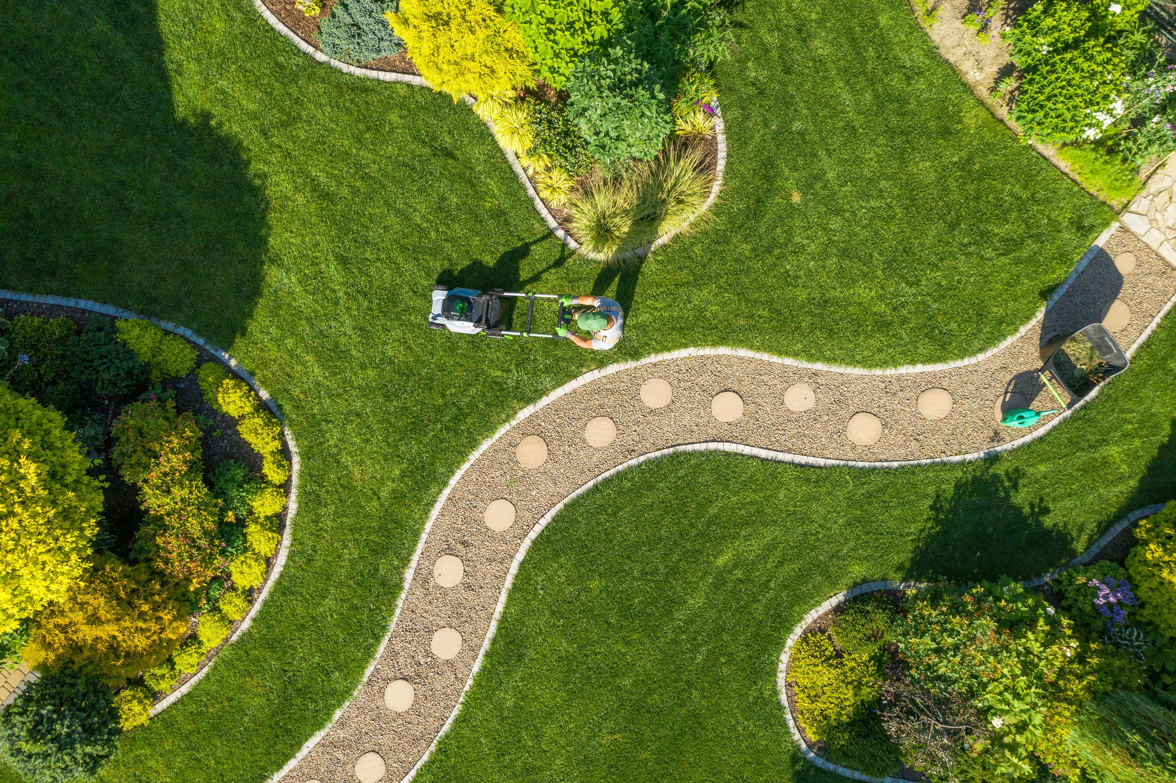 Gemini said Aerial view of a person mowing a green spring yard with a winding gravel path and manicured garden beds.