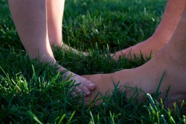Small bare feet standing on bigger bare feet on green grass in lawn