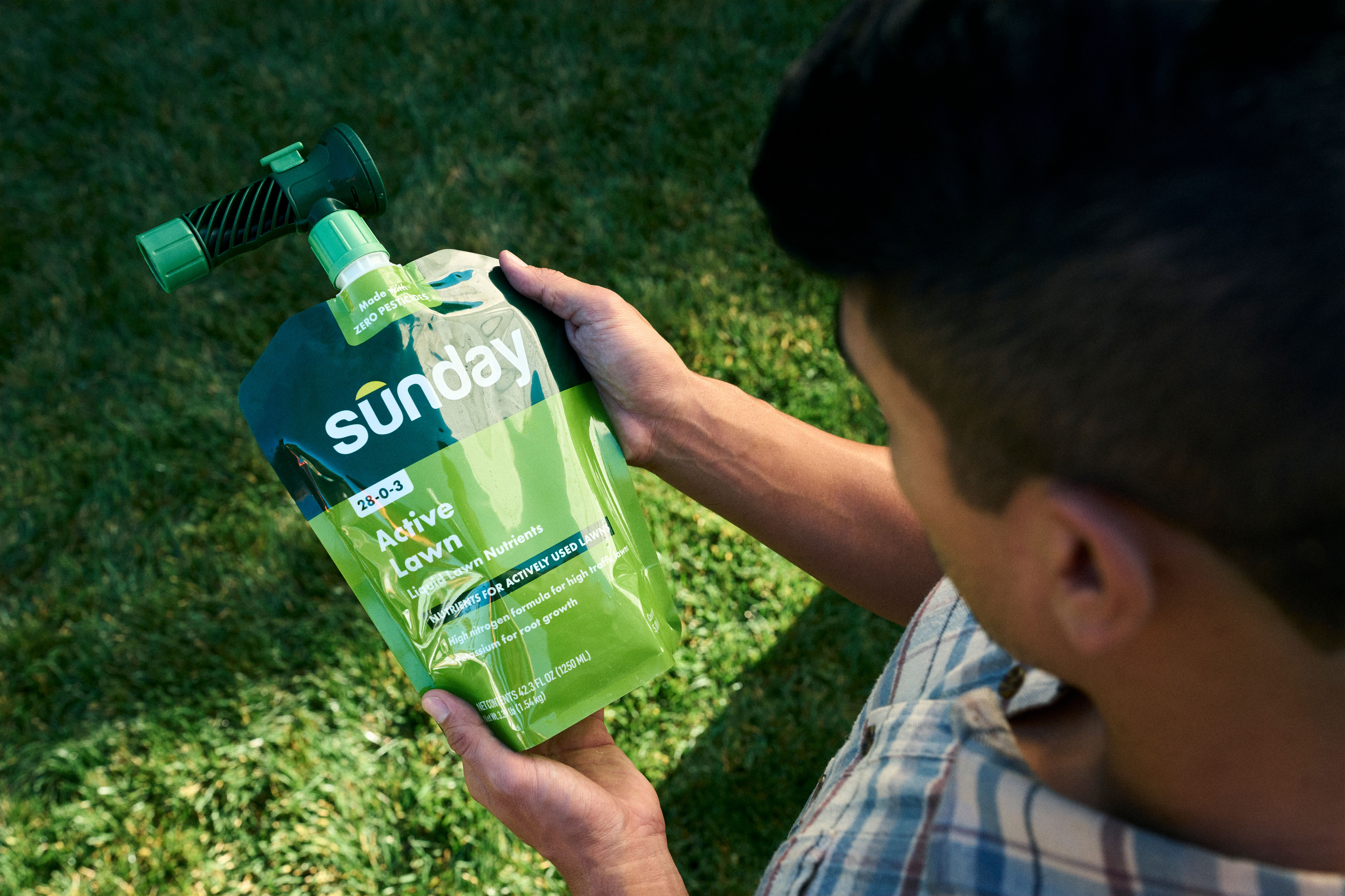 Person holding a Sunday Active Lawn liquid fertilizer pouch with a hose end sprayer attached, standing on a green lawn.