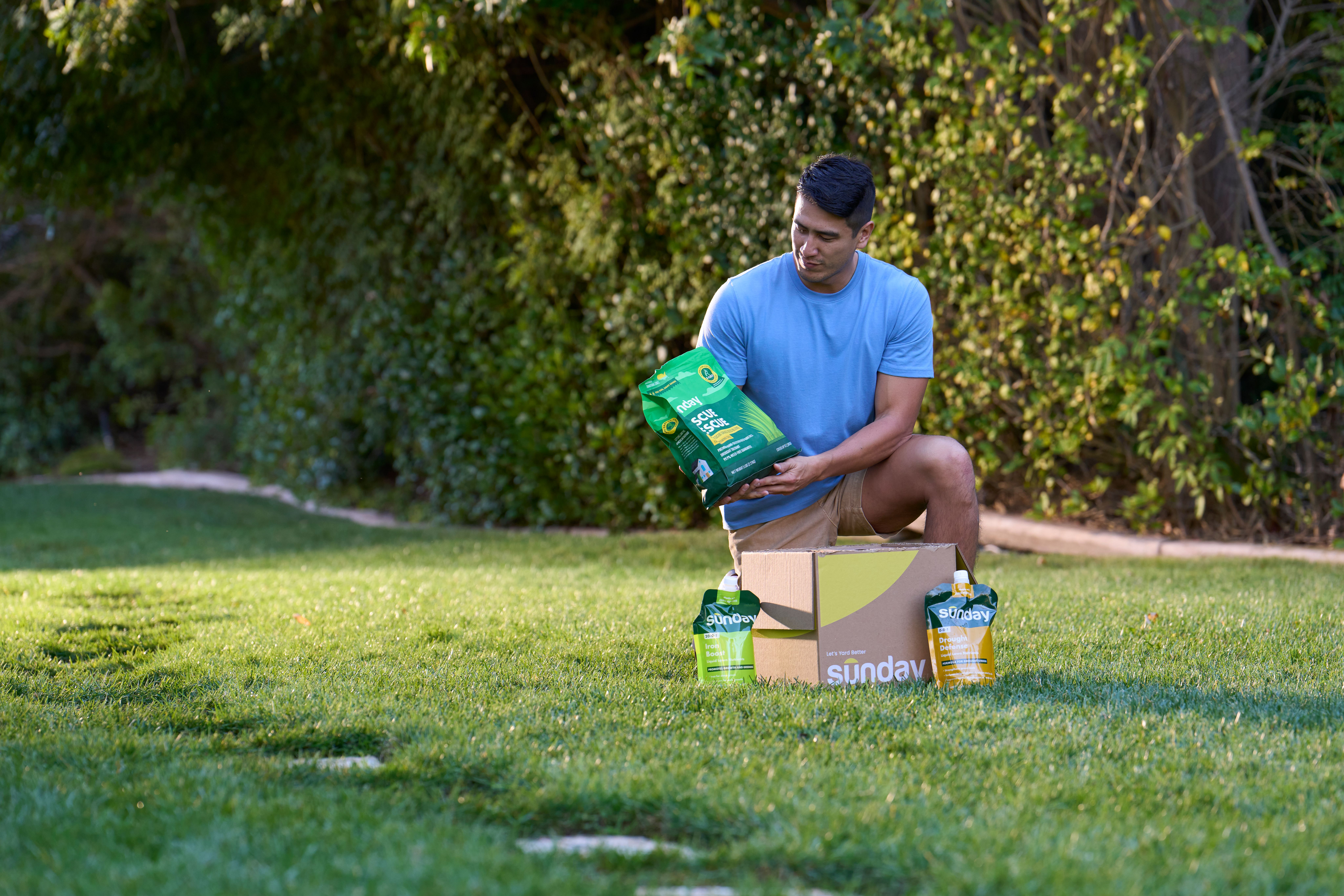 Person kneeling on a lawn beside an open Sunday lawn care box, holding a bag of lawn treatment with Sunday pouches set on the grass.