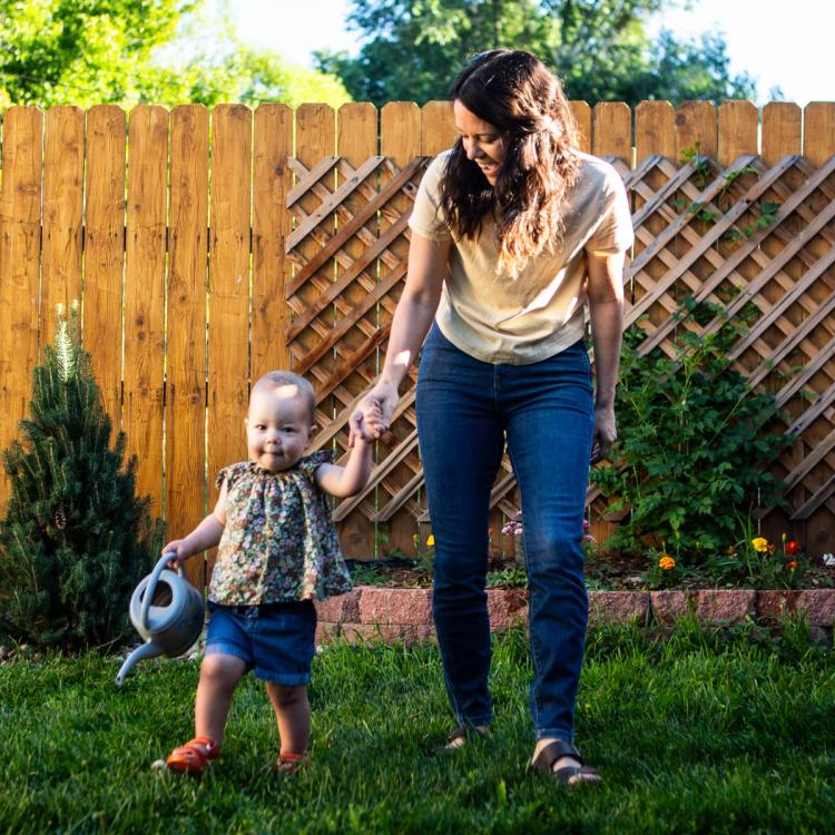 A woman and her toddler walking on a green lawn in a lush garden