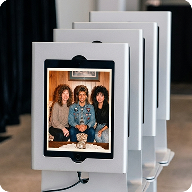 A series of iPads on photo booth mounts are facing the camera.  On the first iPad, the Booth.Events app is showing the result from an AI Prompt capture.  The guests have been transformed into a 90's era basement with wood panelling, big hair and lots of denim.