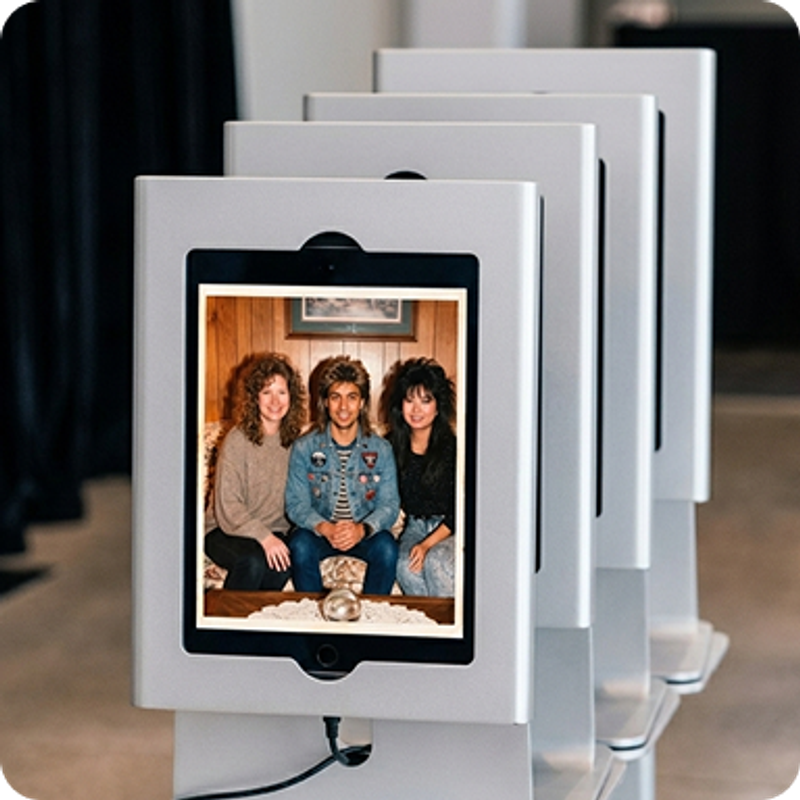 A series of iPads on photo booth mounts are facing the camera.  On the first iPad, the Booth.Events app is showing the result from an AI Prompt capture.  The guests have been transformed into a 90's era basement with wood panelling, big hair and lots of denim.