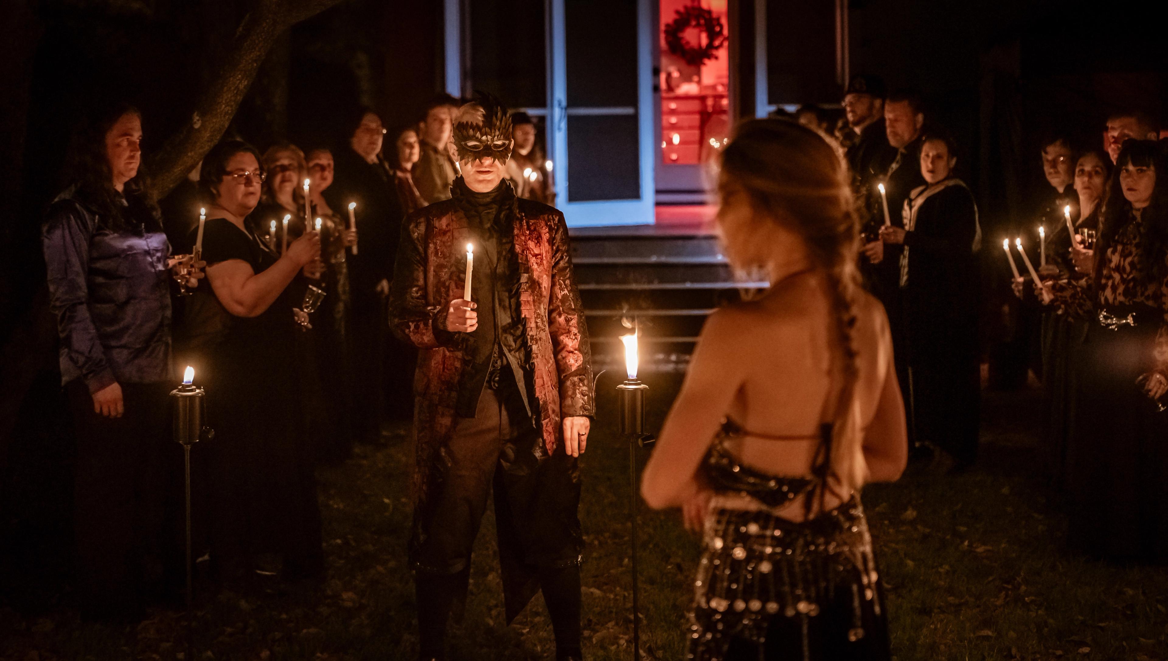 A woman stands with her back to the camera while two rows of people await her, holding candles.