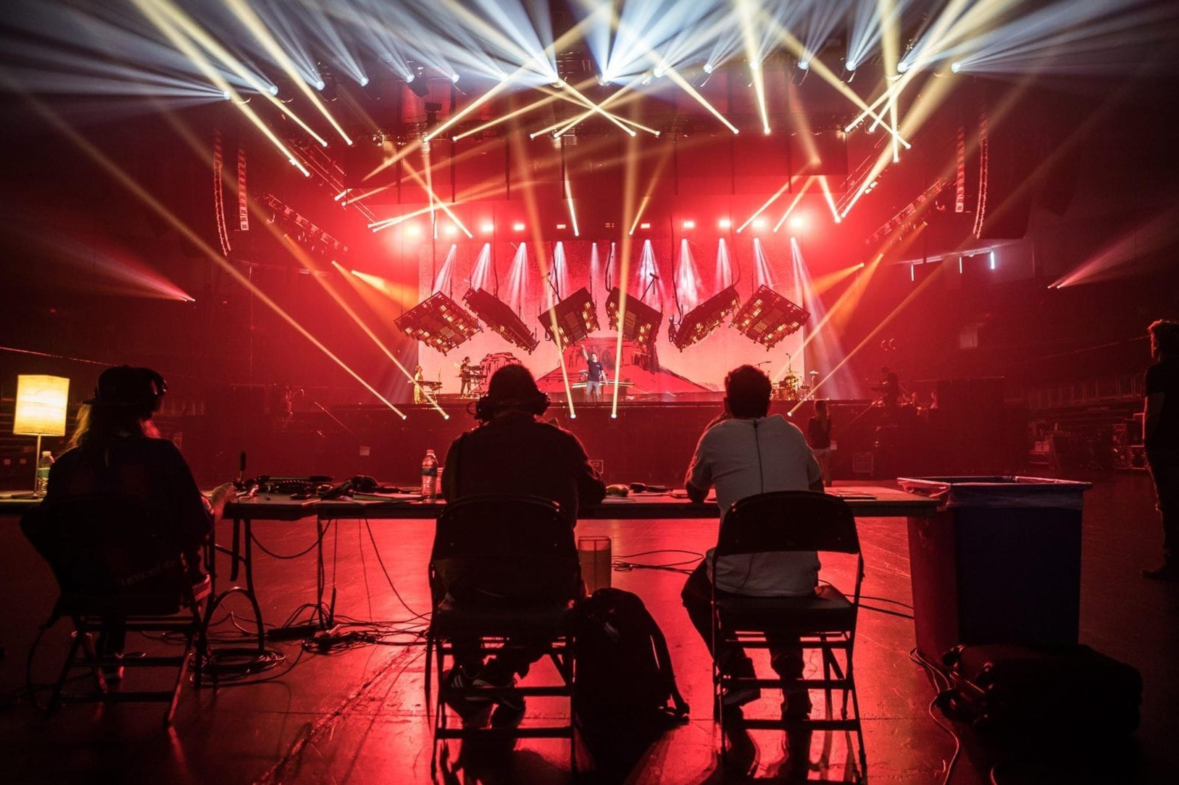 A few working figures sit and stand facing a stage, observing the progress of rehearsals for The Chainsmokers' upcoming tour.