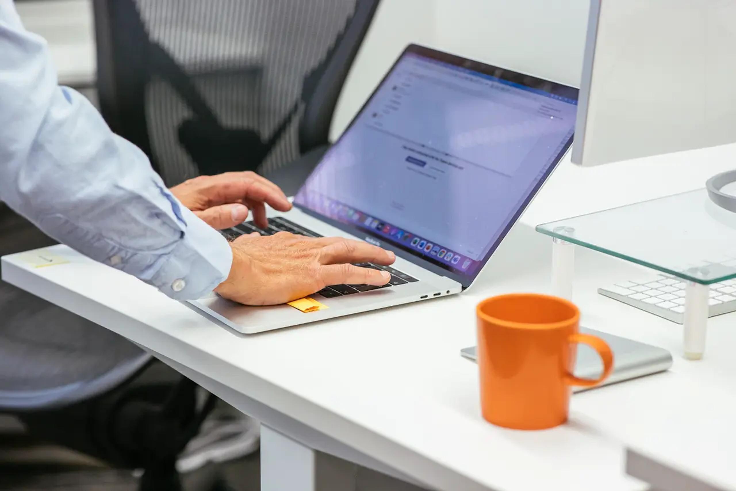 Person typing on a laptop at a white desk next to an orange mug