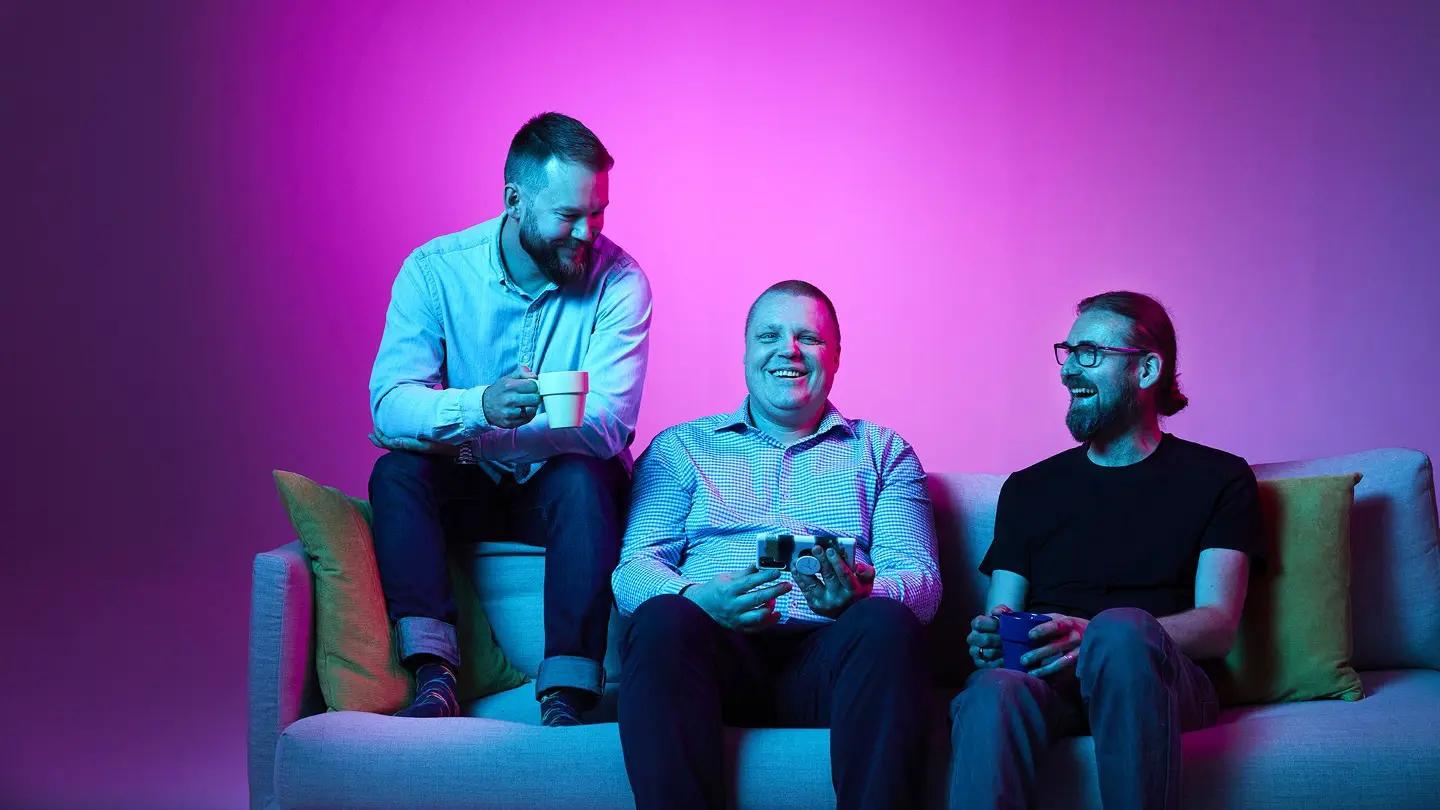 Three men laughing on a sofa against a bright magenta background, holding mugs and a game controller