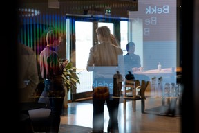 Personer står og snakker sammen i et kontorlokale, sett gjennom en glassvegg med refleksjoner. I bakgrunnen sitter en person ved et møtebord.