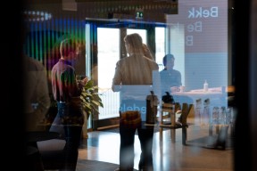 Personer står og snakker sammen i et kontorlokale, sett gjennom en glassvegg med refleksjoner. I bakgrunnen sitter en person ved et møtebord.
