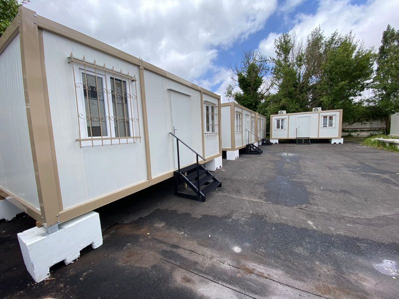 Row of three beige and white container office units on a paved yard in Spain, each raised on footings with its own steps.