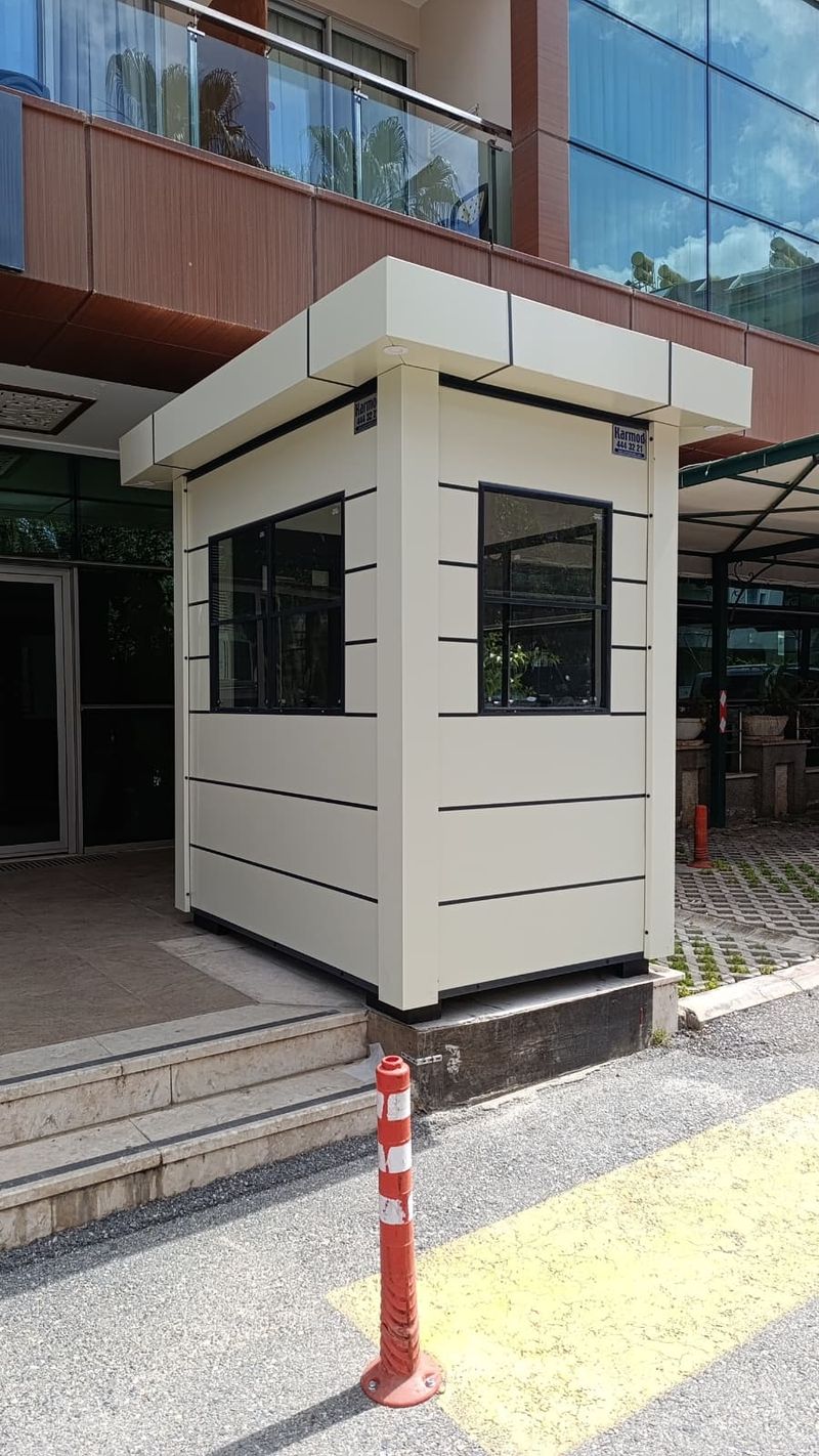 Cream composite-clad gatehouse cabin with a deep overhanging roof, standing at the entrance of a modern office building.