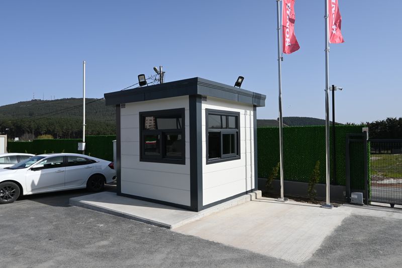 White and anthracite Metro City security cabin beside the vehicle gate of a car showroom, with floodlights and cameras mounted on the roof.