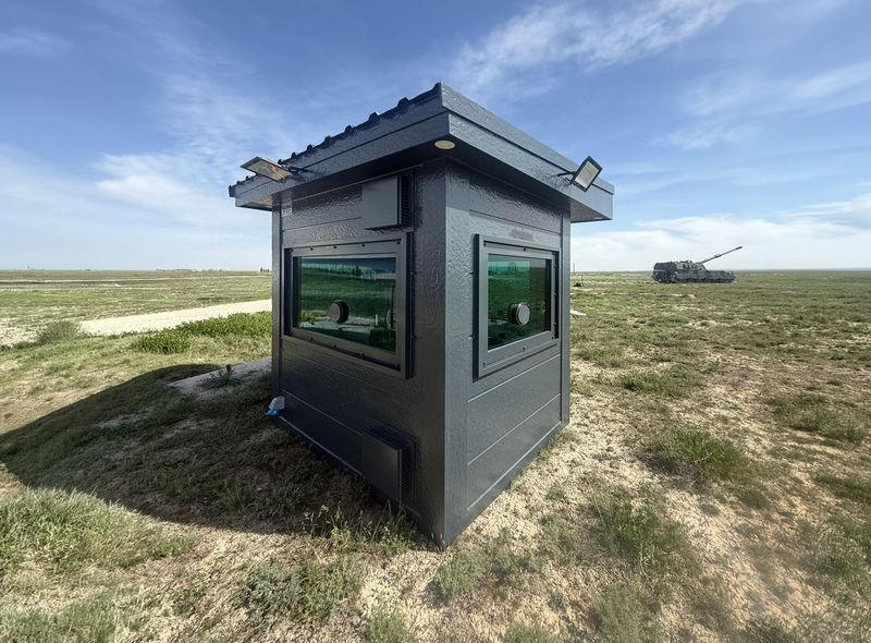 Dark textured armoured cabin with ballistic glazing and firing ports, standing alone on open grassland.