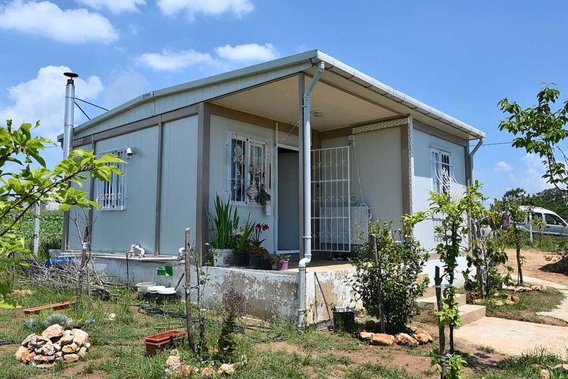 Container home with a canopied porch, a flue and grilled windows on a rural smallholding, with young fruit trees around it.