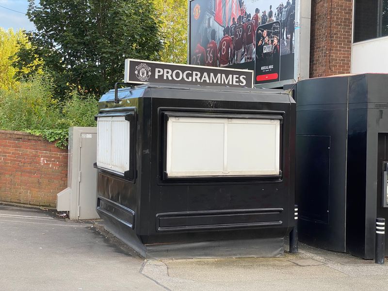 Black GRP Manchester United programme kiosk on the approach to Old Trafford, with a club billboard behind it.