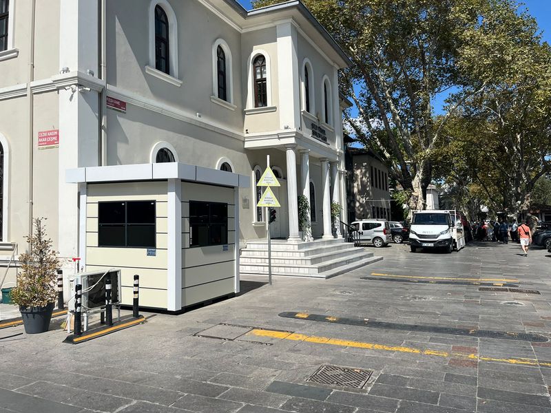 Cream and white composite cabin with a wide roof canopy, set on paving beside a historic foundation building on an Istanbul street.