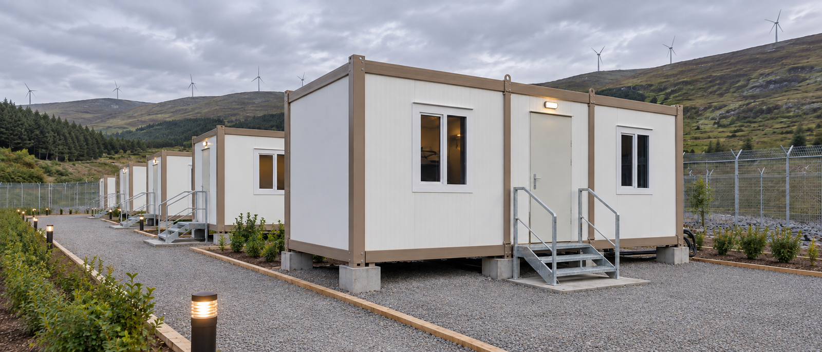 Row of portable accommodation cabins on a gravel site with wind turbines on the hills behind