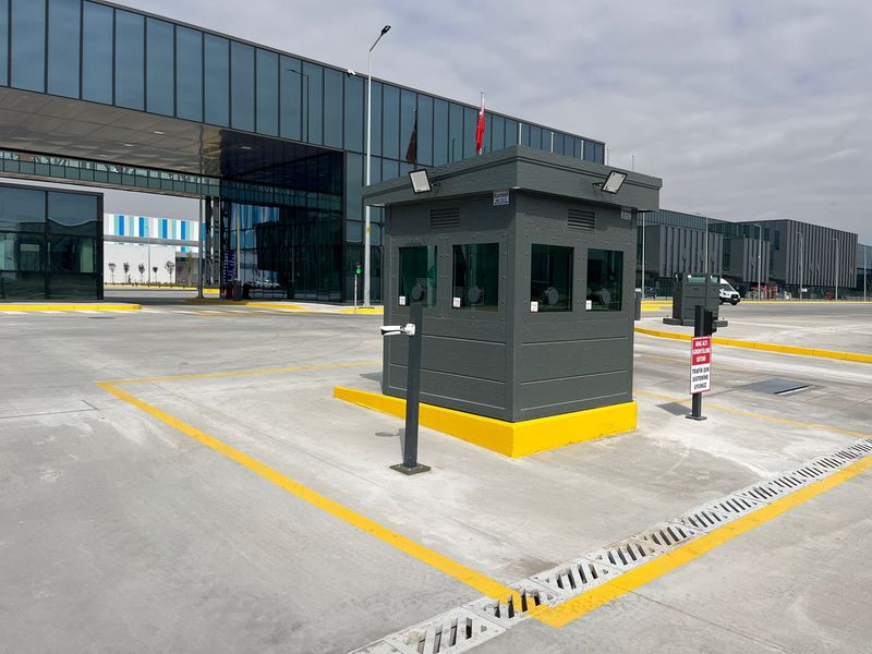 Dark grey armoured security cabin on a yellow-painted plinth, controlling the vehicle gate of a large manufacturing plant.