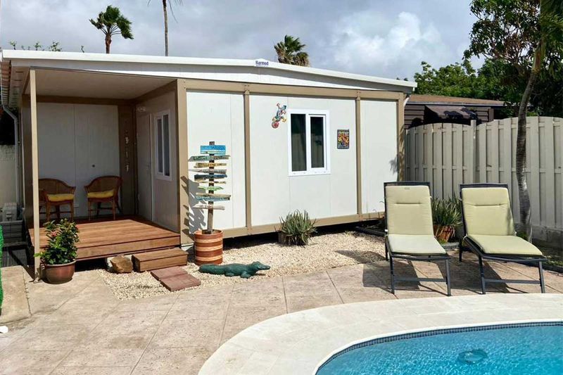 Container guest suite with a timber deck and a covered seating area beside a swimming pool, with palm trees behind.
