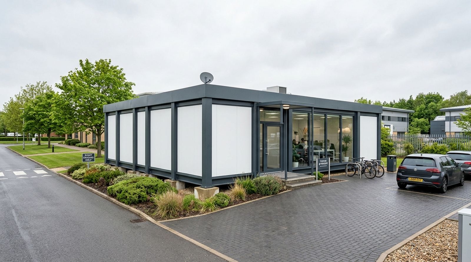 Grey single-storey modular office building with a glazed entrance beside parking bays on a business park