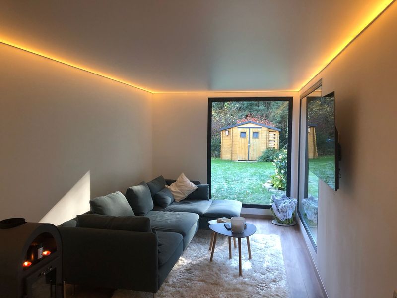 Container house living room lit by concealed cove lighting, with a corner sofa and a full-height window onto the garden.
