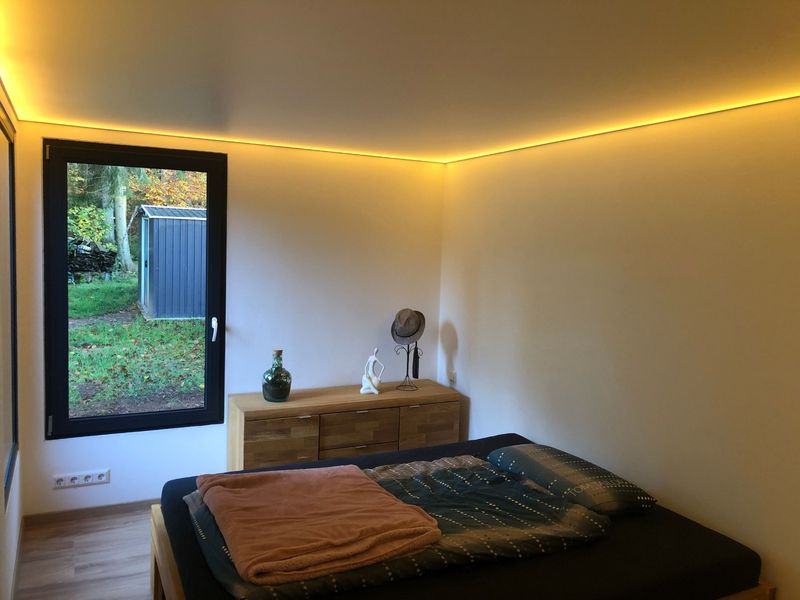 Container house bedroom with concealed cove lighting, a double bed and an oak sideboard, the window looking onto autumn woodland.