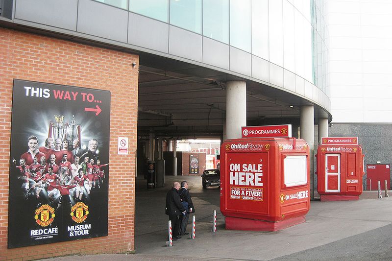 Two red United Review programme kiosks in the entrance concourse at Old Trafford, beneath Manchester United signage.