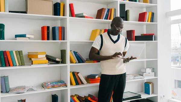 Black man standing in front of bookcase browing the internet on his laptop