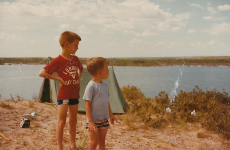 Two boys stand outside a tent at a campground beside a lake  