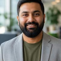 Max, travel virtual assistant, smiling with a full dark beard in a grey blazer over an olive t-shirt