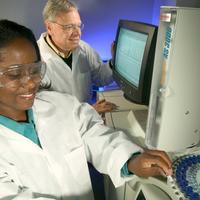 A laboratory technician loads samples into a gas chromatograph while a chemist examines mass-spectral data.