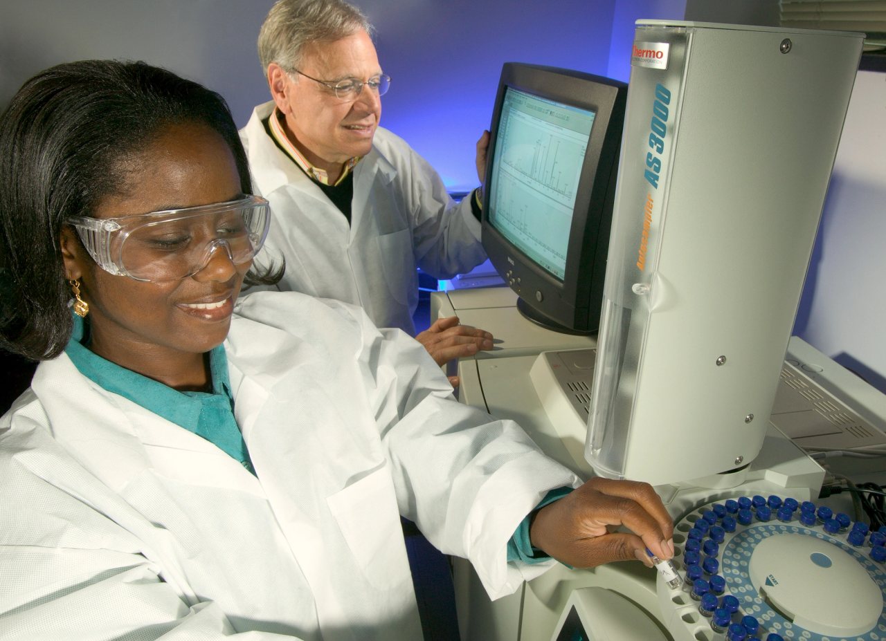 A laboratory technician loads samples into a gas chromatograph while a chemist examines mass-spectral data.