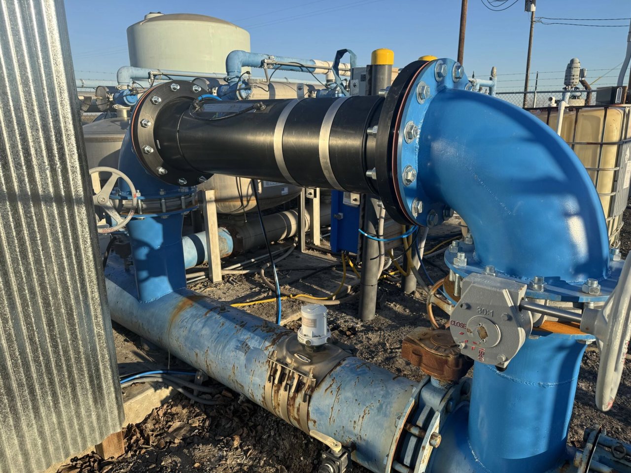 Outdoor agricultural irrigation installation with blue pipe elbows, a black inline unit, valves, and storage tanks.