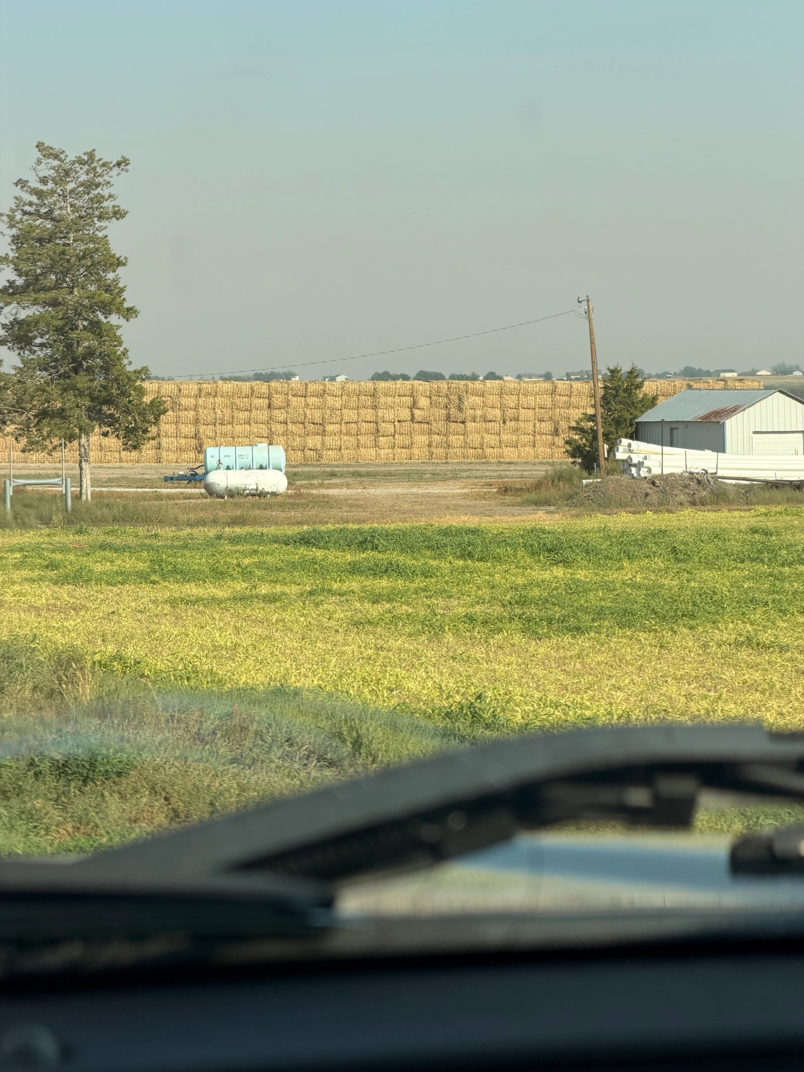 Long stacks of rectangular hay bales beside farm buildings, seen through the windshield.