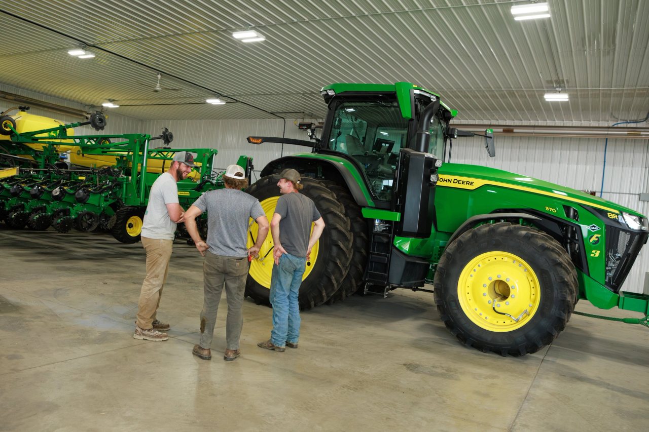 Three people talking beside a tractor and planting equipment inside a farm building.