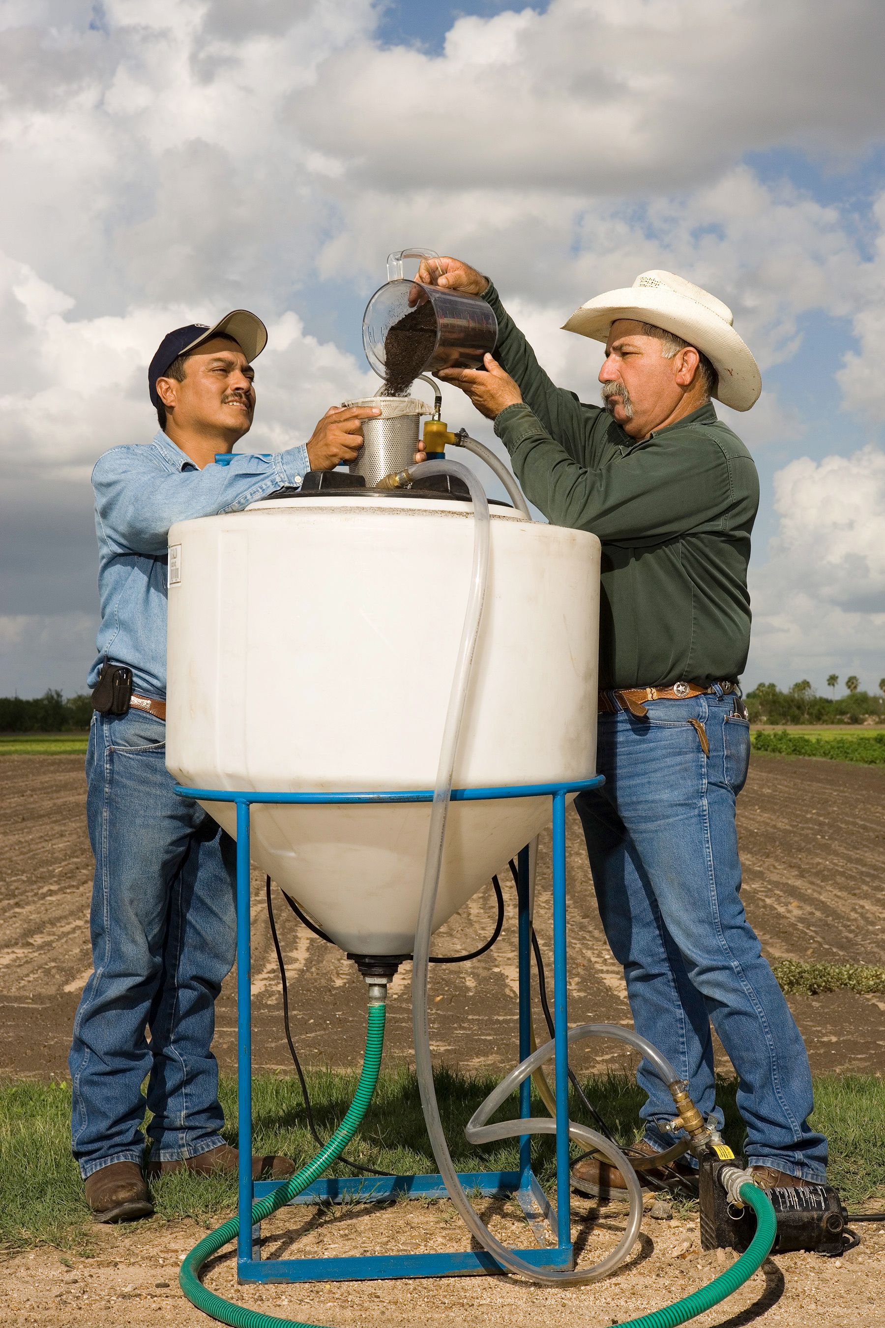 Two workers pour compost into a metal extractor mounted above a large compost tea tank outdoors.