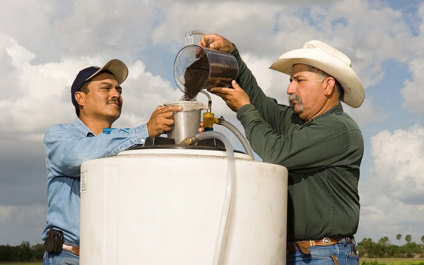 Two workers pour compost into a metal extractor mounted above a large compost tea tank outdoors.