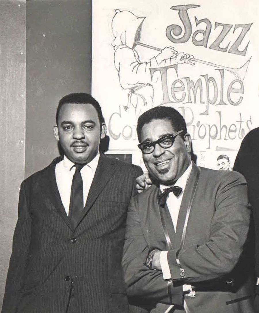 two black men in suits in front of a banner with a hooded figure playing trumpet, reading "Jazz Temple: Co[covered] Prophets"