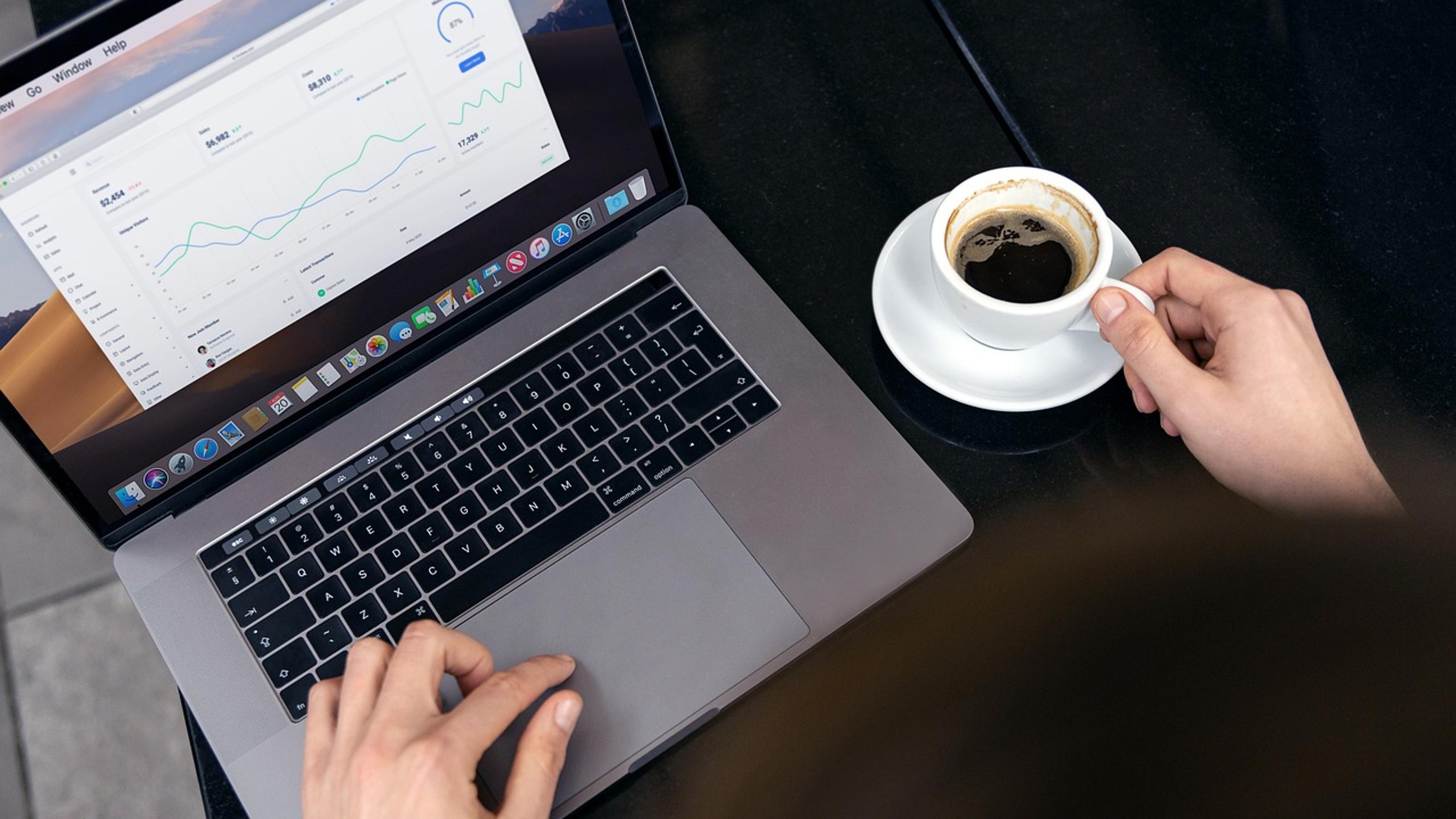 Un professionnel consulte des courbes de chiffres sur son ordinateur portable, une tasse de café à la main.