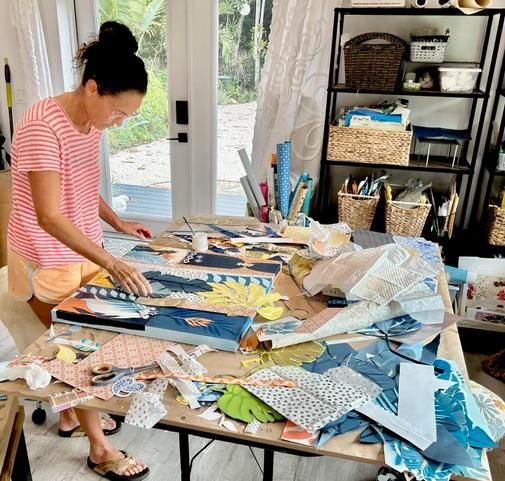 Jennifer arranging paper scraps across canvases on her studio worktable, baskets and shelves behind her by the garden door