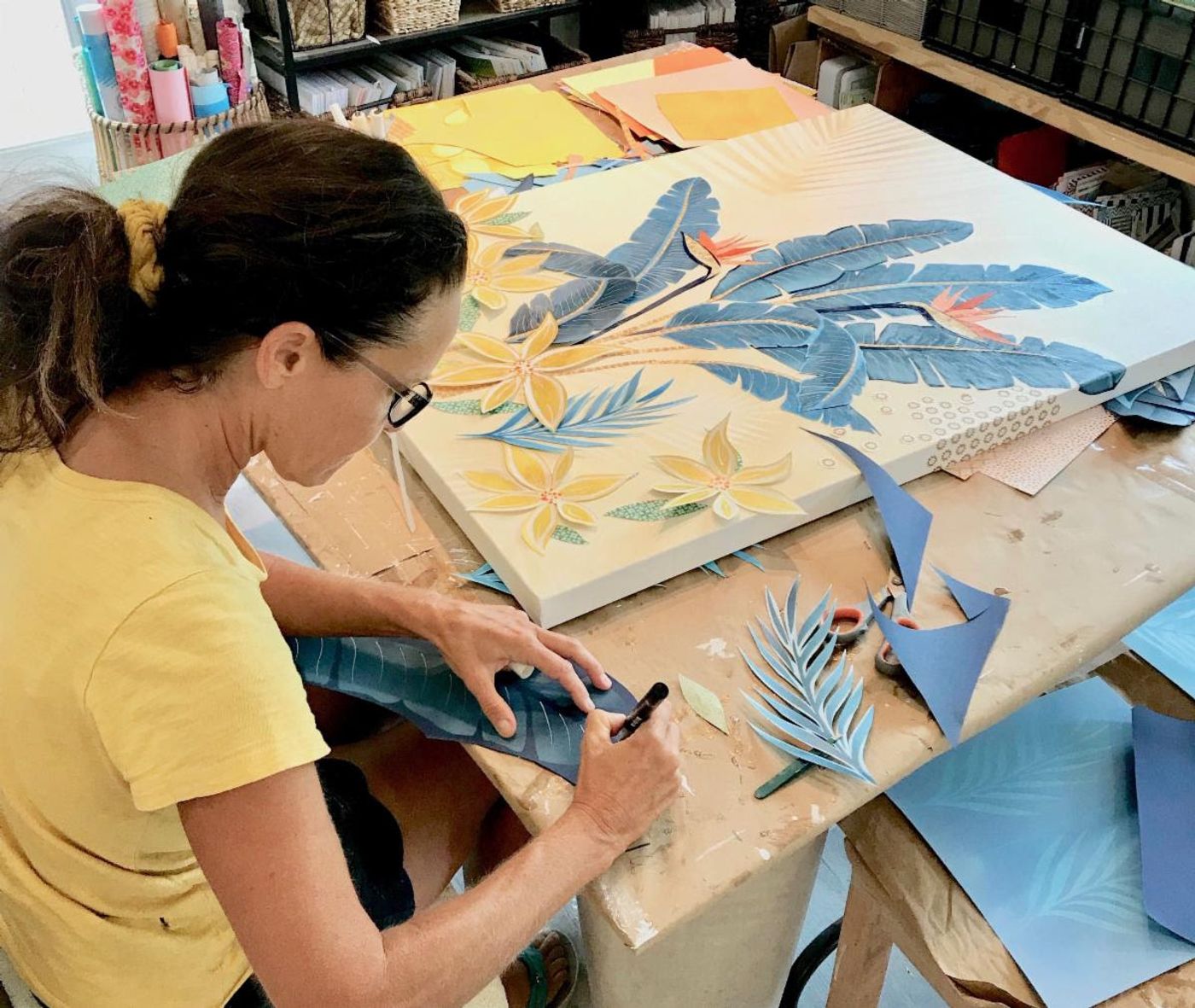 Jennifer draws vein lines on a blue paper leaf beside a nearly finished botanical collage