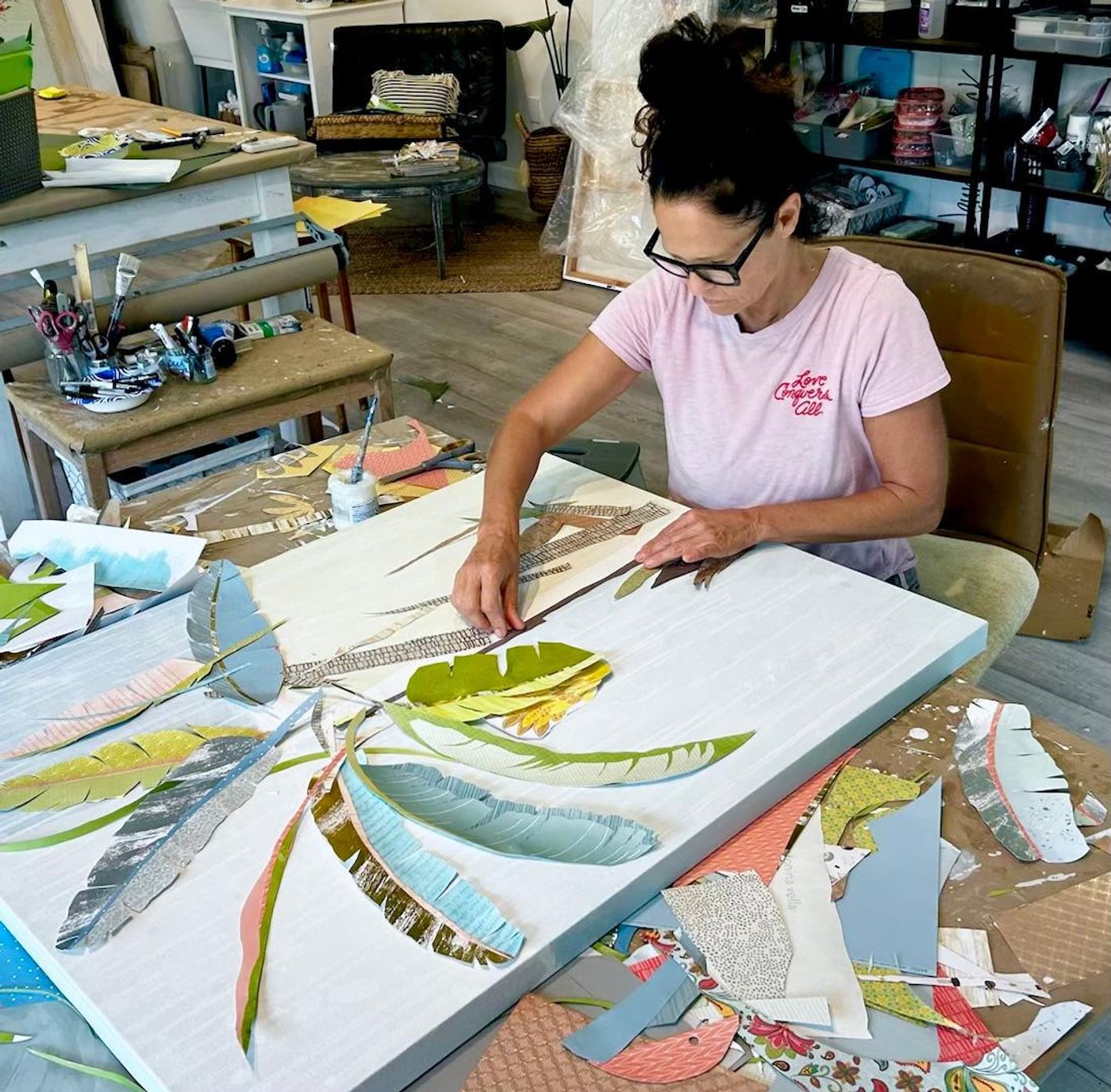 Jennifer presses a brick-patterned paper trunk onto a white canvas, cut banana leaves in sage and blue fanned across the table