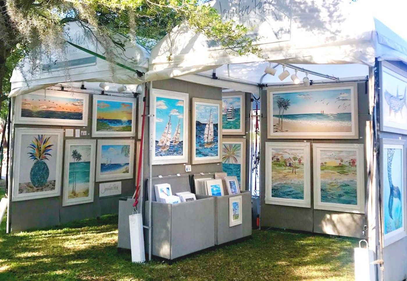 Jennifer's white festival tent hung with framed coastal collages — sailboats, beaches, a blue pineapple — print bins at the center.