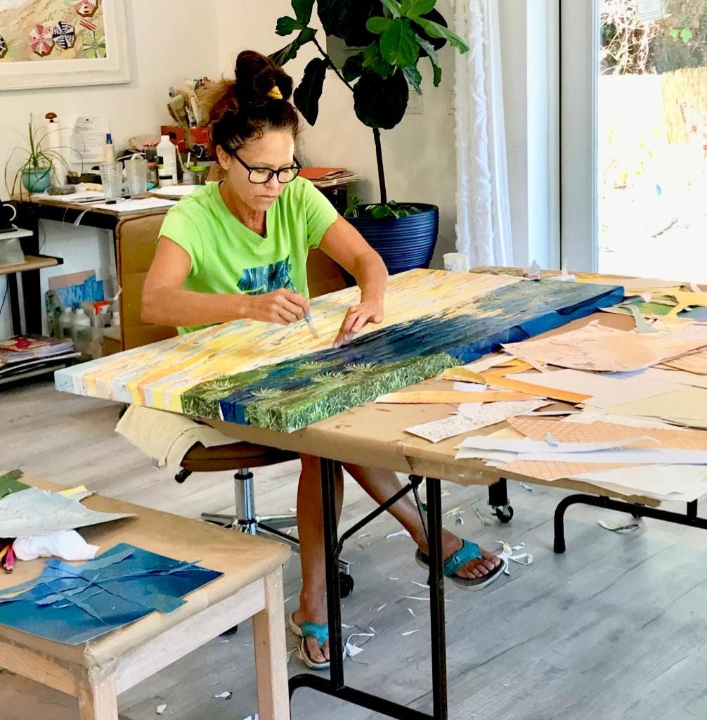 Jennifer seated at her studio table, brush in hand, working across the long bay collage, paper scraps scattered on the floor.