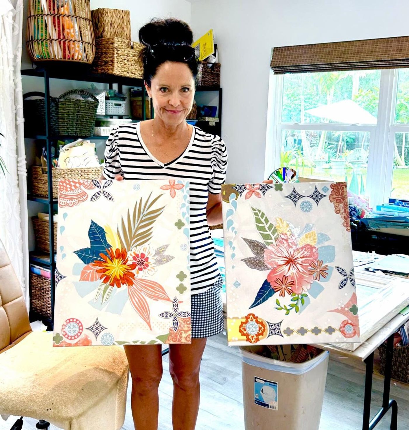 Jennifer in the studio holding the two Rosey Posey collages side by side, baskets of sorted papers shelved behind her