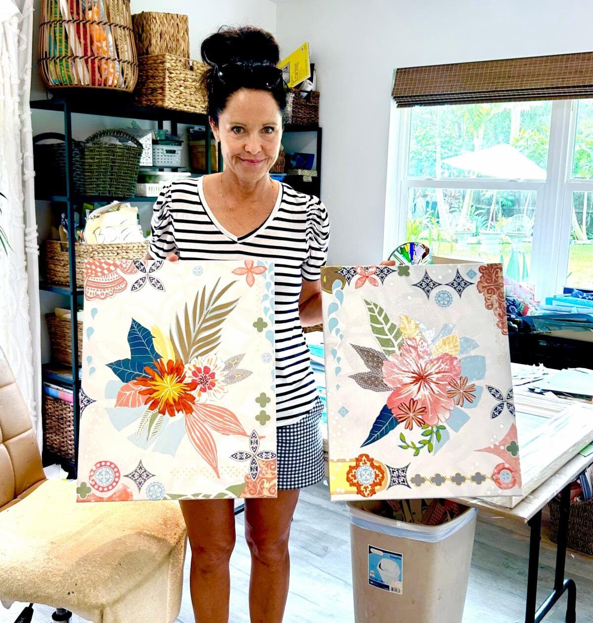 Jennifer in the studio holding the two Rosey Posey collages side by side, baskets of sorted papers shelved behind her